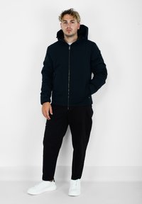 Giacca nera con cappuccio e zip frontale, abbinata a pantaloni neri a sigaretta e sneakers bianche. Design semplice, texture liscia e senza motivi visibili.
