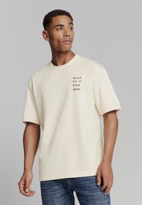 Crèmekleurig katoenen T-shirt met een oversized pasvorm, ronde halslijn, korte mouwen en een textuele afbeelding in bruin op de linkerborst met de tekst "Road To Freedom."