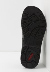 Svart gummisula med texturerade mönster, med räfflor och en röd accent där varumärket "Rieker" är tydligt framhävt.