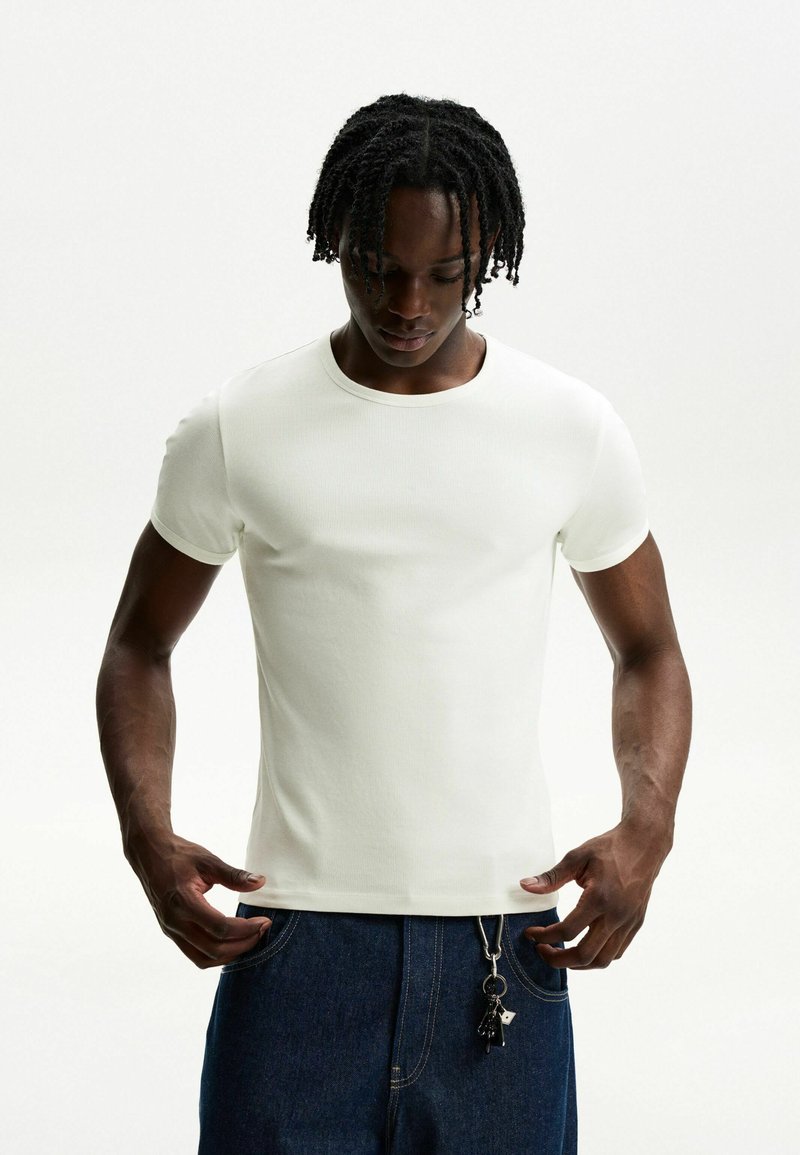 White short-sleeve t-shirt made of ribbed fabric; features a round neckline and a fitted style, paired with dark denim jeans and a keychain.