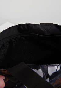 Musta kangaskassi, jonka sisällä on vetoketjullinen tasku, kuviollinen vuori ja osittain näkyvä kukkakuosisesti ulkopuoli mustalla hihnalla.