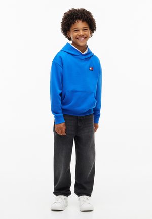 Smilende barn med krøllet hår iført en lys blå hættetrøje, sorte jeans og hvide sneakers, stående foran en ensfarvet hvid baggrund.