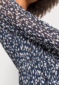 Granatowa sukienka z długimi, transparentnymi rękawami z wzorem w kropki w białym i beżowym kolorze. Materiał wydaje się lekki i teksturowany, z subtelnymi pliskami.