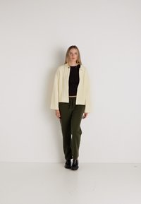 Kremowa kurtka o fakturze z opadającymi ramionami, czarny krótki top oraz oliwkowe spodnie. Całość dopełniają czarne buty. Proste tło.