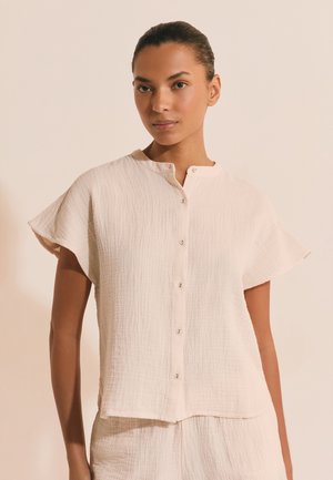 Kobieta w kremowej, teksturowanej bluzce na krótki rękaw, zapinanej na guziki, z okrągłym dekoltem oraz pasujących luźnych spodniach, stojąca na jednolitym tle.