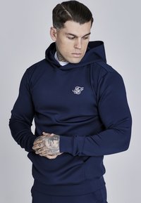Felpa con cappuccio blu navale realizzata in tessuto morbido, con un piccolo logo bianco sul petto, maniche aderenti e tasca a marsupio.