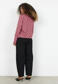 Dlhý rukáv s červeným vzorom a geometrickým dizajnom, doplnený voľnými čiernymi nohavicami a čiernymi lodičkami na podpätku. Jednofarebné pozadie, neutrálny podklad.