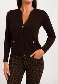 Cardigan marrone scuro con scollatura a V, due tasche frontali e dettagli in bottoni argentati. Indossato su pantaloni a stampa leopardata. Texture a maglia liscia.