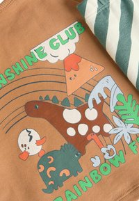 Ruskea puuvillapaita, jossa on leikkisiä grafiikoita dinosauruksesta, tulivuoresta ja eläimistä kirkkaissa väreissä, sekä teksti "SUNSHINE CLUB". Vihreät sävyviivat.