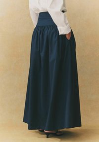 Jupe maxi plissée marine avec une taille haute et des poches latérales, en tissu lisse. Associée à une chemise blanche à boutons et des talons noirs.