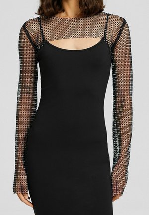 Mujer que lleva un vestido negro con corpiño ajustado y mangas largas de malla transparente con un patrón de diamantes y pequeños adornos.