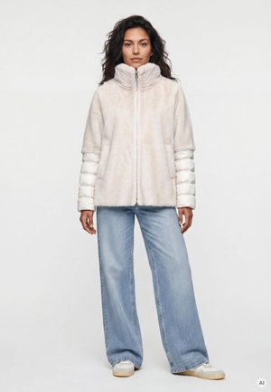 Frau steht und trägt eine hellbeige Zip-up-Jacke mit gesteppte Ärmeln, weitgeschnittene hellblaue Jeans und weiße Sneaker vor einfachem Hintergrund.