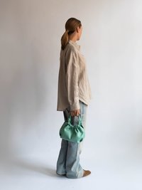 Zelena torba z gladko teksturo, v kombinaciji z svetlo modrimi flare jeans hlačami in bež lanenim srajcem, model obrnil se vstran.