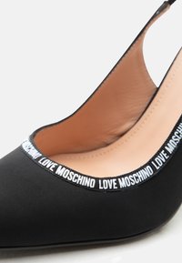 Μαύρο γυναικείο παπούτσι με ψηλό τακούνι και μυτερή μύτη, με μπεζ εσωτερικό και λευκή επιγραφή «LOVE MOSCHINO» κατά μήκος του περιγράμματος του ανοίγματος του παπουτσιού.