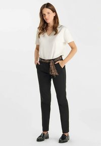 Witte V-hals T-shirt gecombineerd met zwarte broek. De broek heeft een riem met luipaardprint. Model draagt zwarte loafers en staat met handen in de zakken.