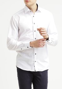 White long-sleeve shirt with a fitted design, subtle texture, and contrasting dark buttons. Cuffs feature similar button accents.