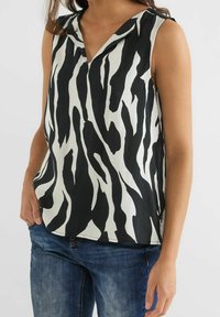 Žena ve bezrukávovém topu s černo-bílým zebrovým vzorem a výstřihem do V, v kombinaci s modrými džíny, stojící na jednobarevném pozadí.