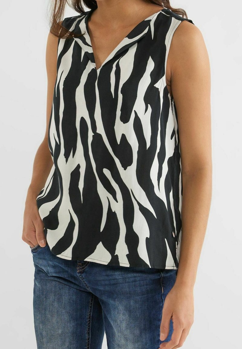 Žena ve bezrukávovém topu s černo-bílým zebrovým vzorem a výstřihem do V, v kombinaci s modrými džíny, stojící na jednobarevném pozadí.