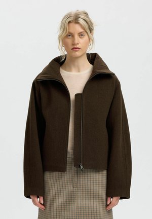 Blond kvinde iført en mørkebrun lynlåsjakke over en beige top og ternet nederdel, stående mod en ensartet lys baggrund.