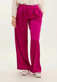 Pantalons larges fuchsia en satin avec des plis, dotés d'une taille haute et d'une fermeture à boutons sur le devant, associés à des baskets blanches.