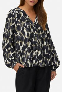 Frau trägt eine langärmlige Bluse mit schwarzen, weißen und olivgrünen abstrakten Tiermotiven und eine schwarze Hose.