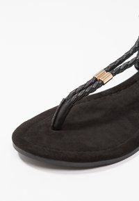 Zwarte teensandalen met gevlochten banden, accenten van goudkleurig beslag en een zachte suède textuur op het voetbed. Plat profiel en minimalistisch ontwerp.