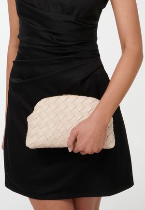 Vævet beige clutch lavet af blødt materiale, med en afrundet top og en subtil glans. Holdes i hånden mod en mørk kjole.