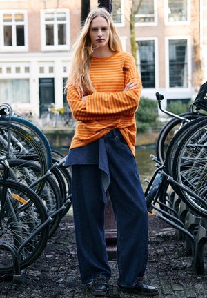 Jonge vrouw met lang blond haar staat met gekruiste armen, draagt een oranje gestreepte trui en brede blauwe spijkerbroek bij geparkeerde fietsen.