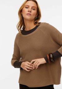 Bruine geribde trui met een ronde hals en lange mouwen. Voorzien van contrasterende donkerbruine manchetten en een houten armband om de pols.