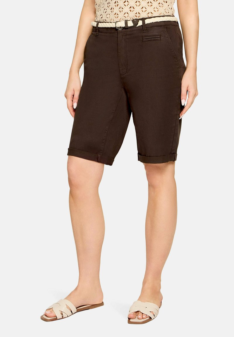 Femme portant un short marron foncé jusqu'aux genoux avec un ourlet roulé, une ceinture tissée beige et des sandales beiges à enfiler, debout sur un fond uni.