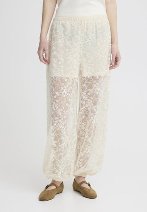 Frau trägt lockere, cremefarbene Spitzenhose mit elastischen Bündchen, kombiniert mit braunen Mary-Jane-Schuhen und einem ärmellosen weißen Oberteil.