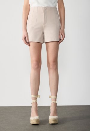 Femme portant un short beige taille haute avec des plis devant et des espadrilles compensées beiges avec des liens à la cheville sur un sol gris.