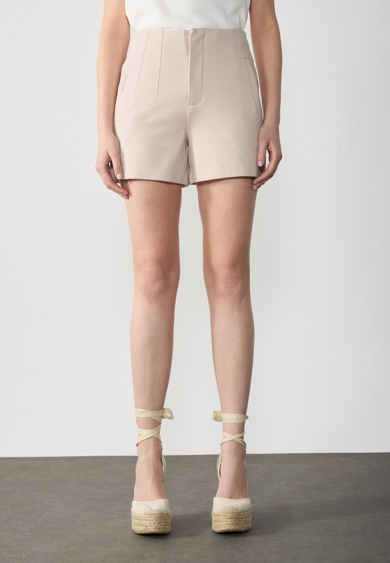 Femme portant un short beige taille haute avec des plis devant et des espadrilles compensées beiges avec des liens à la cheville sur un sol gris.