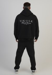 Melna kapuce ar baltu "SIKSILK" logotipu aizmugurē, saskaņota ar melnām sporta biksēm. Audums izskatās mīksts, ar brīvu piegriezumu.