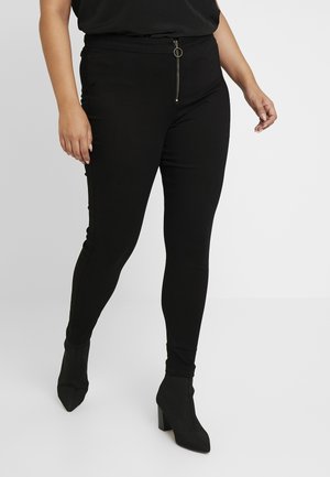 Woman wearing black fitted pants with front zipper detail and black heeled ankle boots, standing against a plain light background.