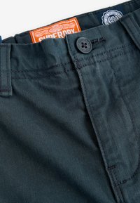 Donkergroene broek met oranje en blauwe merktekens, een zwarte knoopsluiting en een klassieke denimtextuur. Beschikt over stevige stikseldetails.