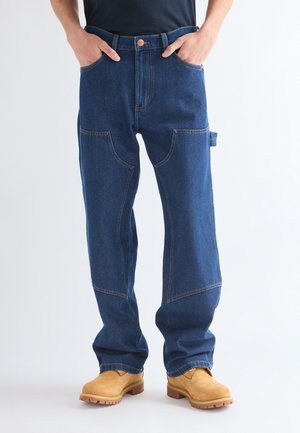 Persoon draagt blauwe denim werkbroek met meerdere zakken en beige werkschoenen, handen in de zakken, staand tegen een effen achtergrond.