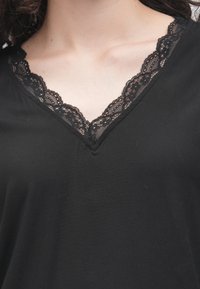 Svart topp med V-utringning pyntet med intrikat svart blonderkant. Mykt stoff, løs passform og subtil drapering.