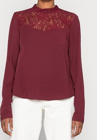 Blusa bordeaux con maniche lunghe e sovrapposizione di pizzo nella parte superiore, caratterizzata da un colletto rotondo e una consistenza liscia.