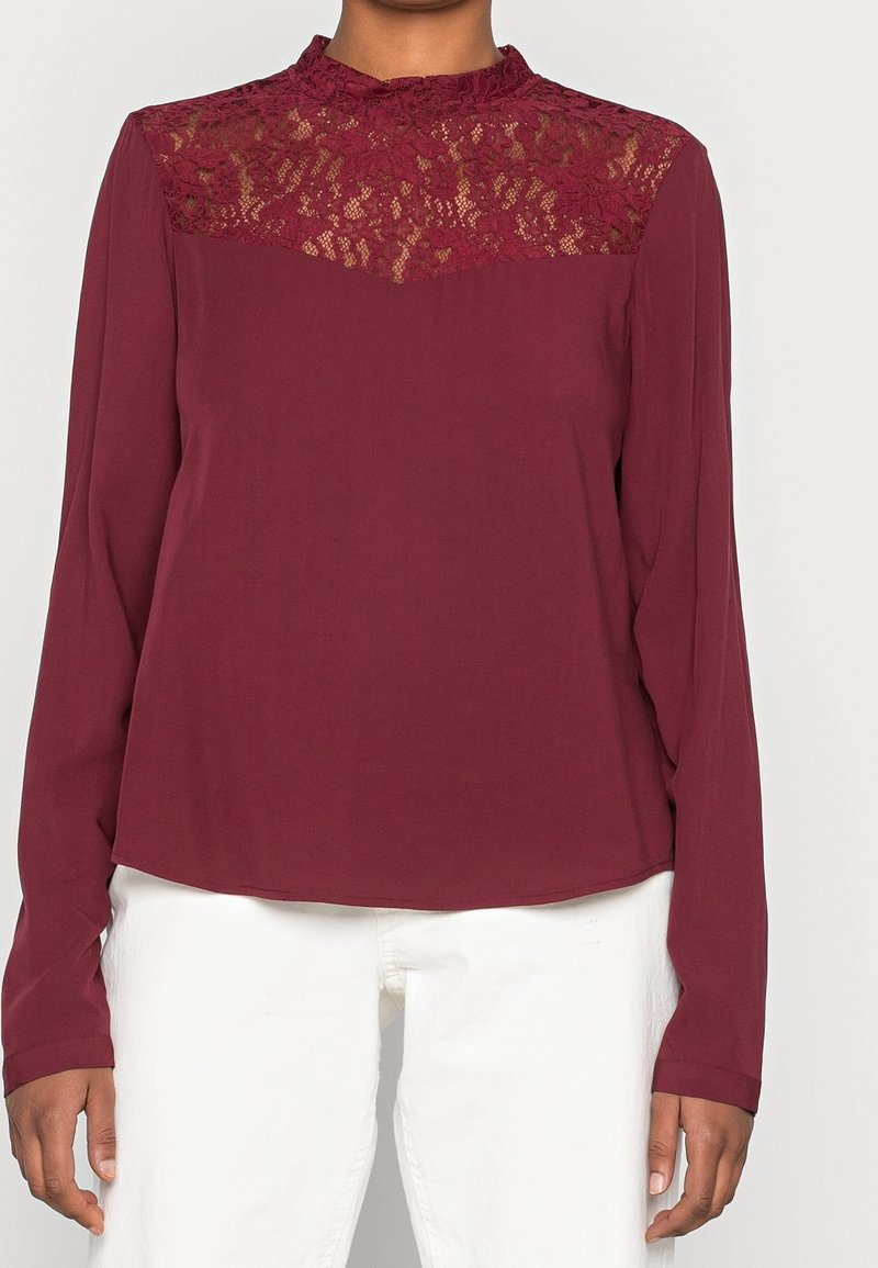 Blusa bordeaux con maniche lunghe e sovrapposizione di pizzo nella parte superiore, caratterizzata da un colletto rotondo e una consistenza liscia.