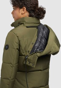 Puffer jacket in olive green with a high collar, zippered pocket, and black inner lining. Features drawstring and snap buttons.