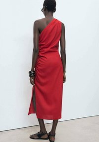 Robe rouge asymétrique avec détail froncé, fente sur le côté, coupe ajustée, tissu fluide, portée avec des sandales noires et des bracelets.