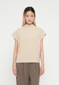 Top beige a maniche corte con collo alto e design morbido, realizzato in tessuto liscio, abbinato a pantaloni marroni larghi.