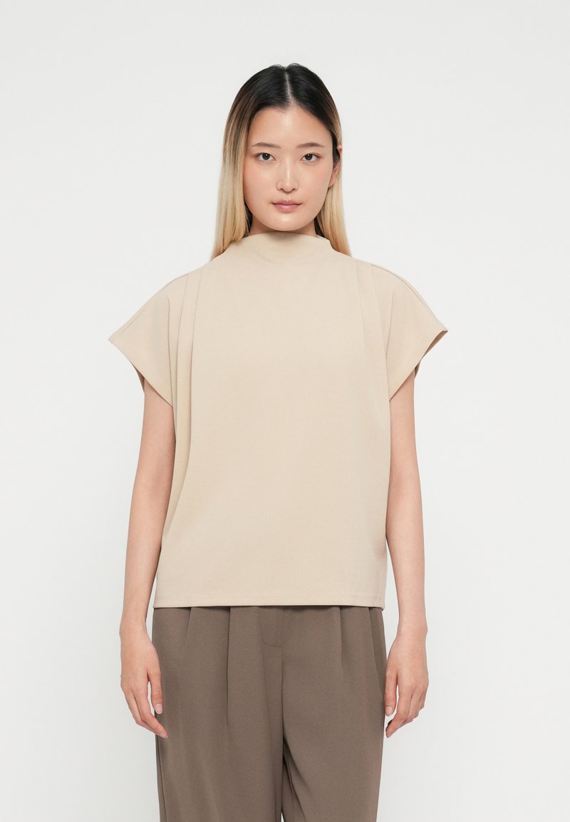 Top beige a maniche corte con collo alto e design morbido, realizzato in tessuto liscio, abbinato a pantaloni marroni larghi.