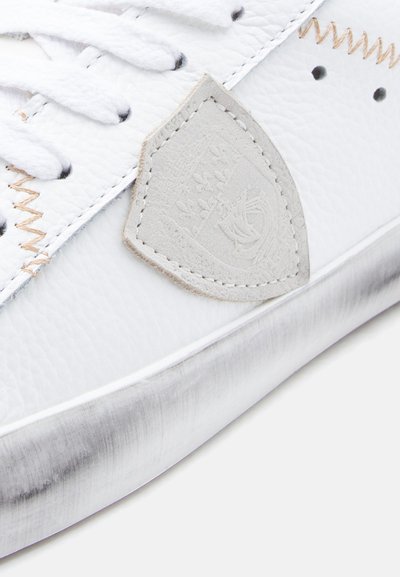 Λευκό δερμάτινο αθλητικό παπούτσι με υφή στην επιφάνεια, αντίθετη ραφή και ανοιχτό γκρι λογότυπο. Διαθέτει ελαφρώς φθαρμένη λαστιχένια σόλα.