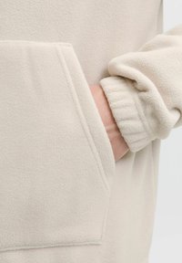 Käsi työnnettynä vaaleanbeige fleece-takin taskuun, jossa elastinen hihna näkyy ranteessa.