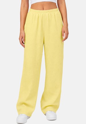 Geel broek met wijde pijpen en elastische taille, gedragen door een persoon met witte sneakers en een wit cropped top, zichtbaar van midriff tot voeten.