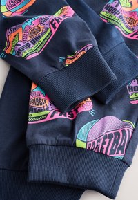 Tamsiai mėlynas džemperis su rankogaliais ir ryškiais grafikos atspaudais rožinės, žalios ir oranžinės spalvų, vaizduojančiais sporto temas ir linksmus piešinius.