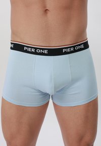 Torso masculino vistiendo bóxers de color azul claro con una cinturilla negra que dice "PIER ONE" sobre un fondo liso.