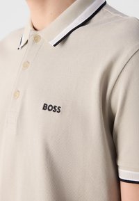 Gros plan d'un polo beige avec trois boutons, col et bords de manches rayés noir et blanc, et « BOSS » brodé sur la poitrine.